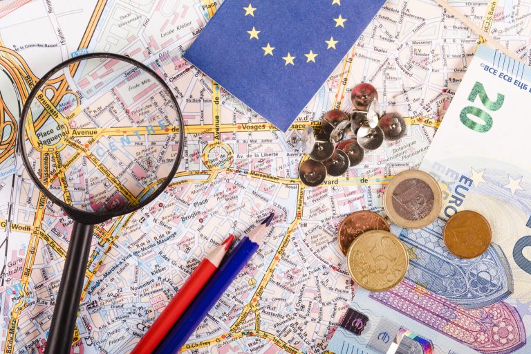 euro-currencies-with-magnifying-glass-pushpins-pencils-flag-world-map-scaled